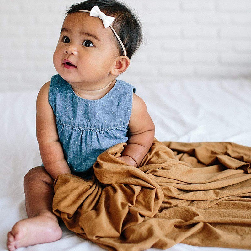 Baby in a blue dress with a white bow sitting on a white blanket with a brown blanket draped over them.