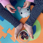 Child's hands holding a small Melli snack container of snacks on a colorful play mat.