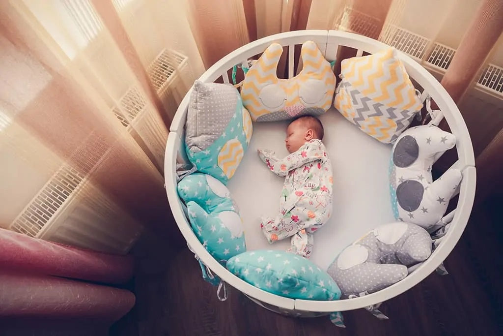 Baby sleeping in a round baby crib with colorful pillows and soft toys.