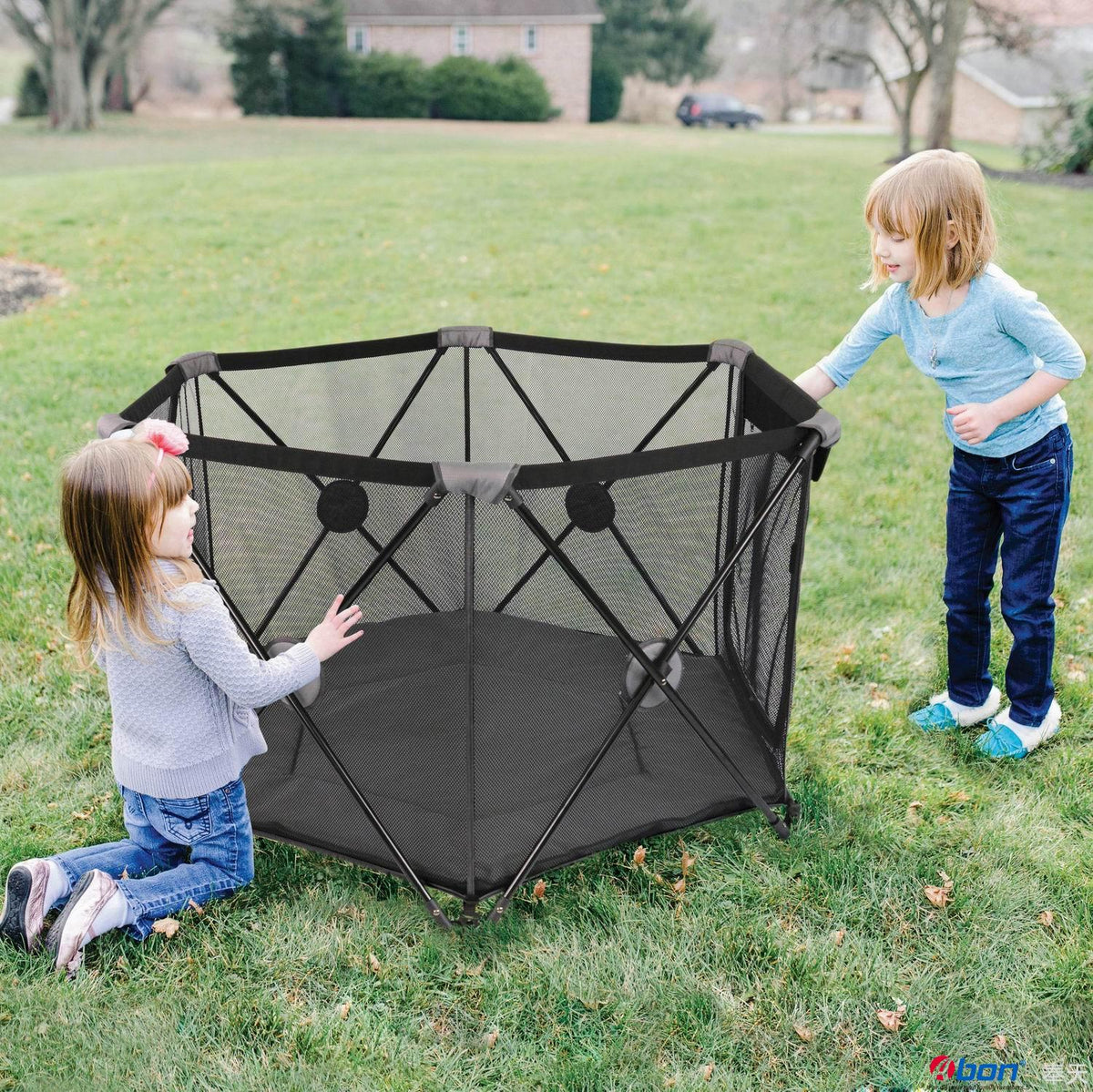 A black mesh hexagonal Magic go playpen is set up on a grassy lawn, with two young children interacting with it. One child is kneeling on the left, and the other is standing on the right.