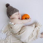 Baby sleeping with a small pumpkin, wearing a knitted hat and wrapped in a beige blanket.