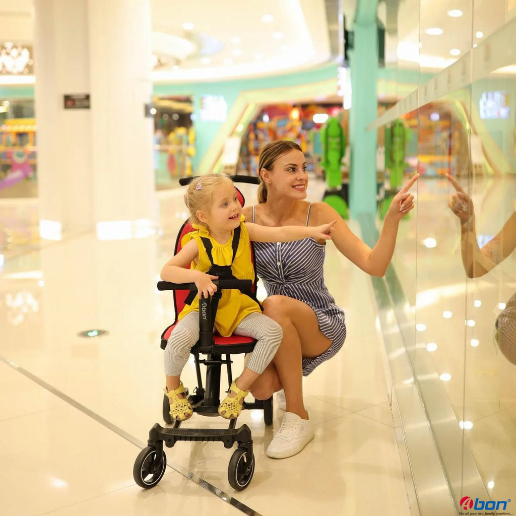 A woman and a young girl in a yellow outfit pose with a black and red convertible rider ll stroller in a bright, modern shopping mall. 