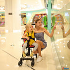 A woman and a young girl in a yellow outfit pose with a black and red convertible rider ll stroller in a bright, modern shopping mall. 