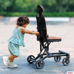 A toddler in a green gingham romper pushes a black and brown convertible rider ll stroller outdoors on a paved walkway.