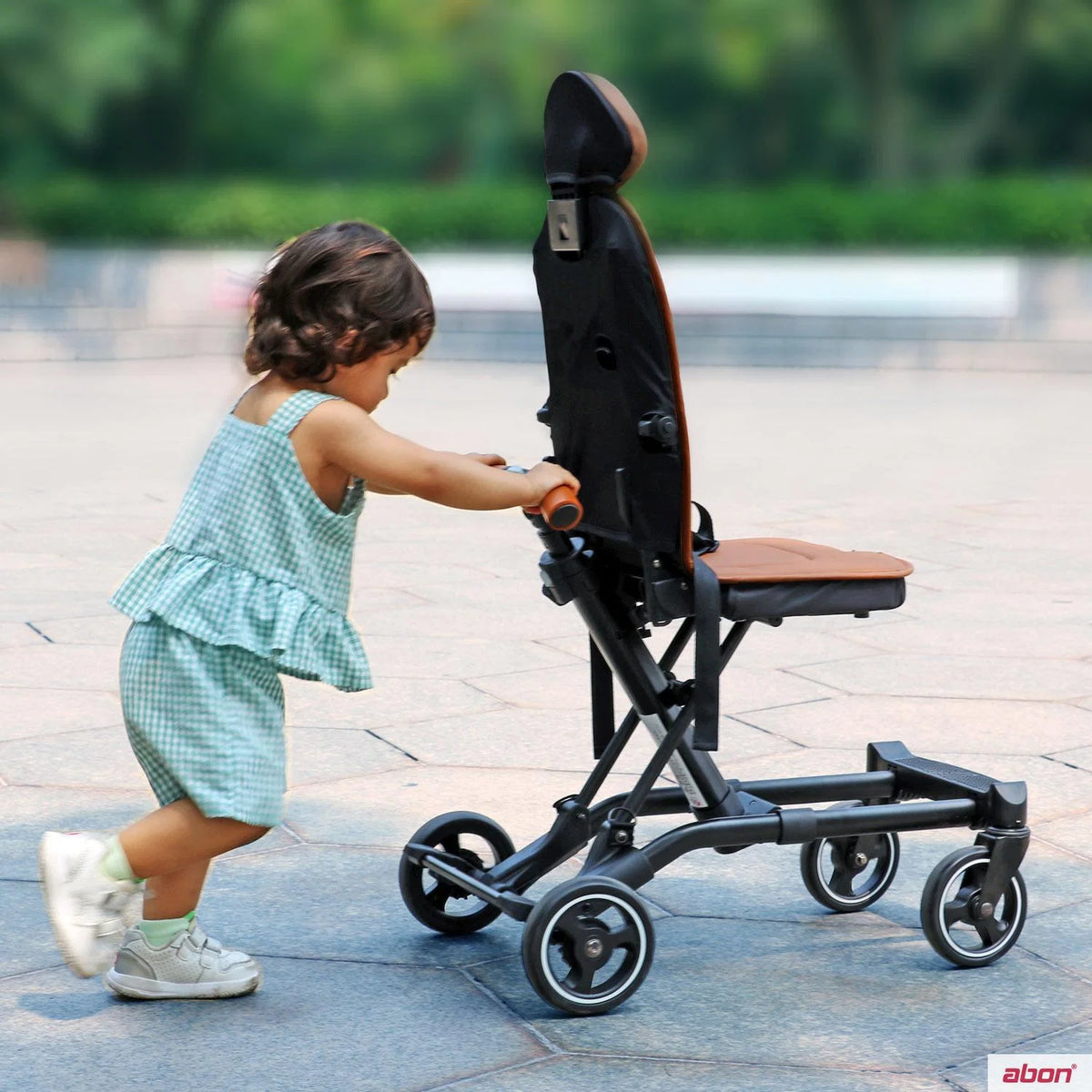 A toddler in a green gingham romper pushes a black and brown convertible rider ll stroller outdoors on a paved walkway.