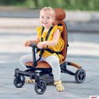 A young child wearing a yellow top and striped leggings sits in a brown and black Convertible rider ll stroller-style tricycle outdoors on a paved surface. The product has a brown seat and backrest, black frame and handlebars, and small black wheels.