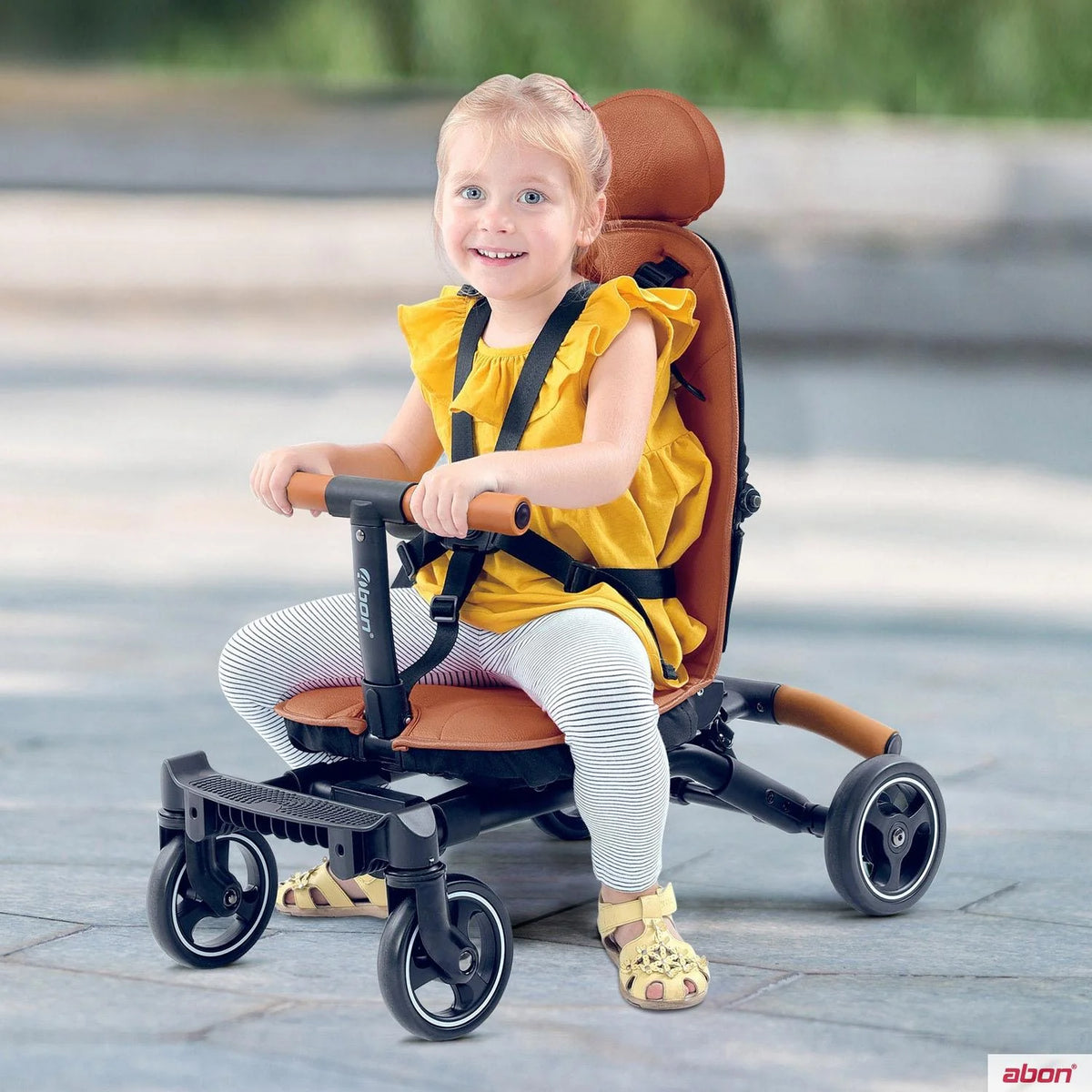 A young child wearing a yellow top and striped leggings sits in a brown and black Convertible rider ll stroller-style tricycle outdoors on a paved surface. The product has a brown seat and backrest, black frame and handlebars, and small black wheels.